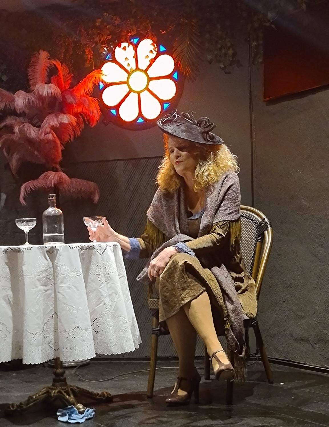 The actress dressed in 1920s clothing and a fascinator, sits at a cafe table drinking absinth.